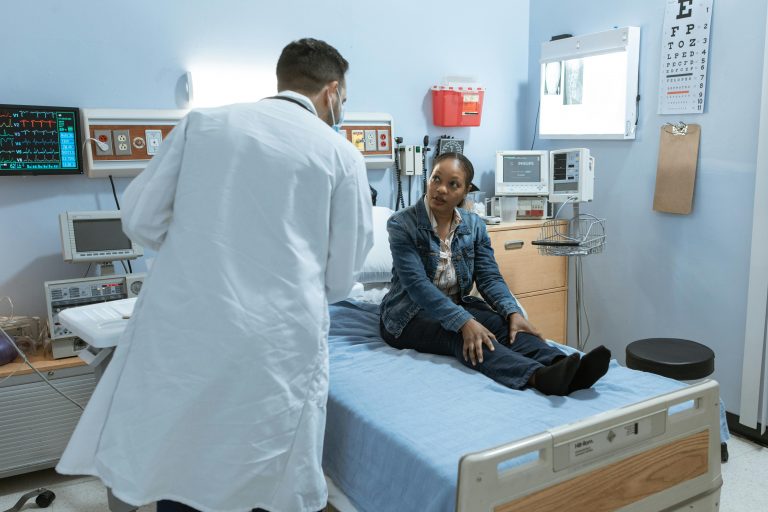 A doctor in a white coat and face mask speaks with a patient sitting on a hospital bed in a medical examination room.