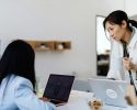 Two women working in an office; one is seated using a laptop while the other stands beside her holding a white mug and looking at a computer screen.