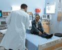 A doctor in a white coat and face mask speaks with a patient sitting on a hospital bed in a medical examination room.